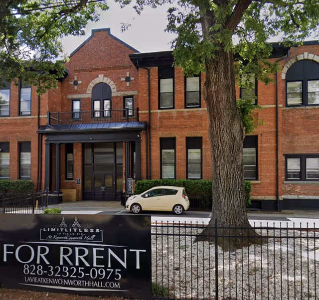 Front facade of a red-brick two-story building with an entrance, a parked car, and a 'For Rent' sign in front.