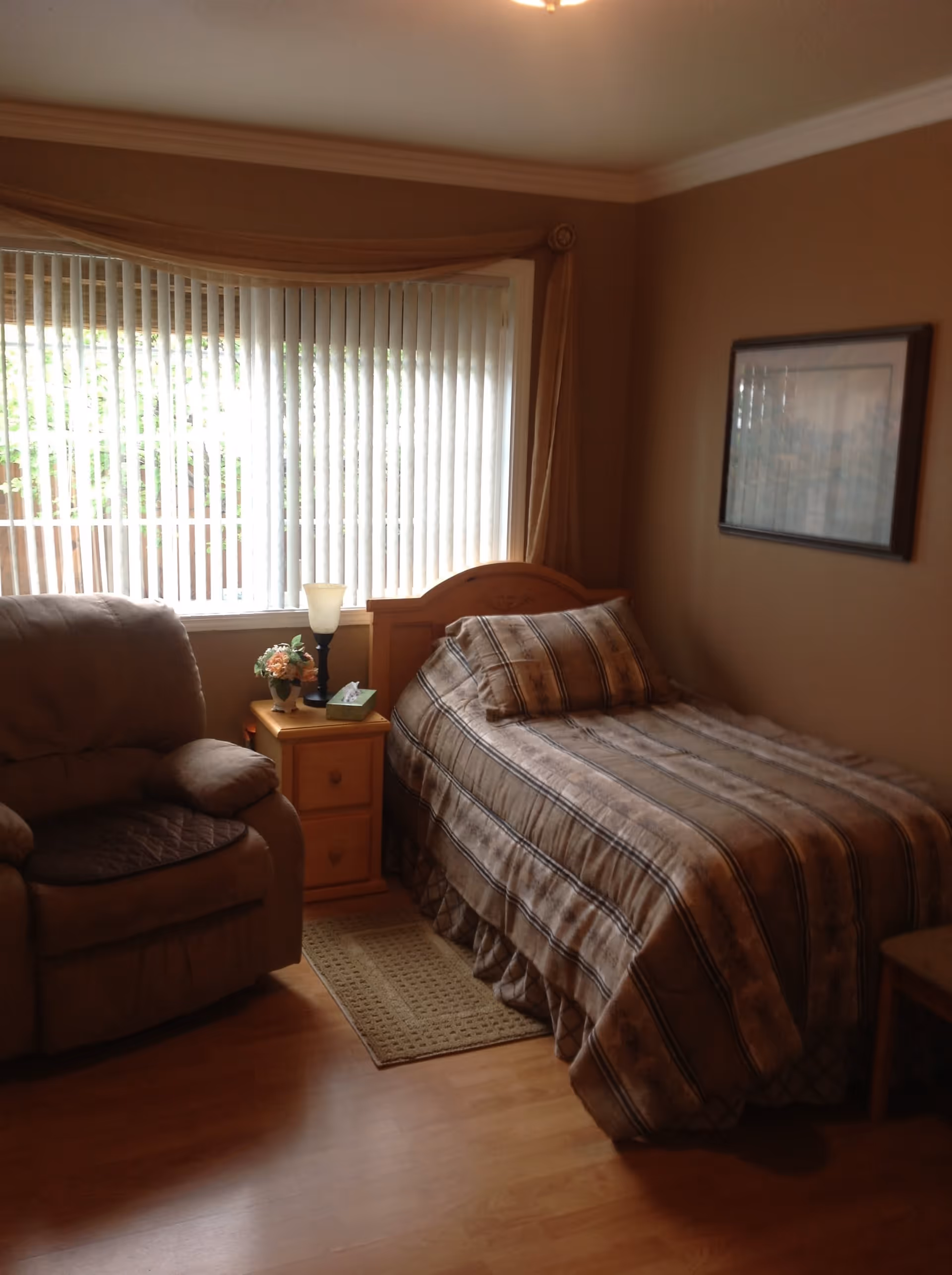 A cozy bedroom with a single bed covered in a striped comforter and matching pillow. Next to the bed is a wooden nightstand with a lamp, a small flower arrangement, and a tissue box. A comfortable armchair is positioned near a large window with vertical blinds and beige curtains. The walls are painted a warm brown color and a framed picture hangs on the wall above the bed.