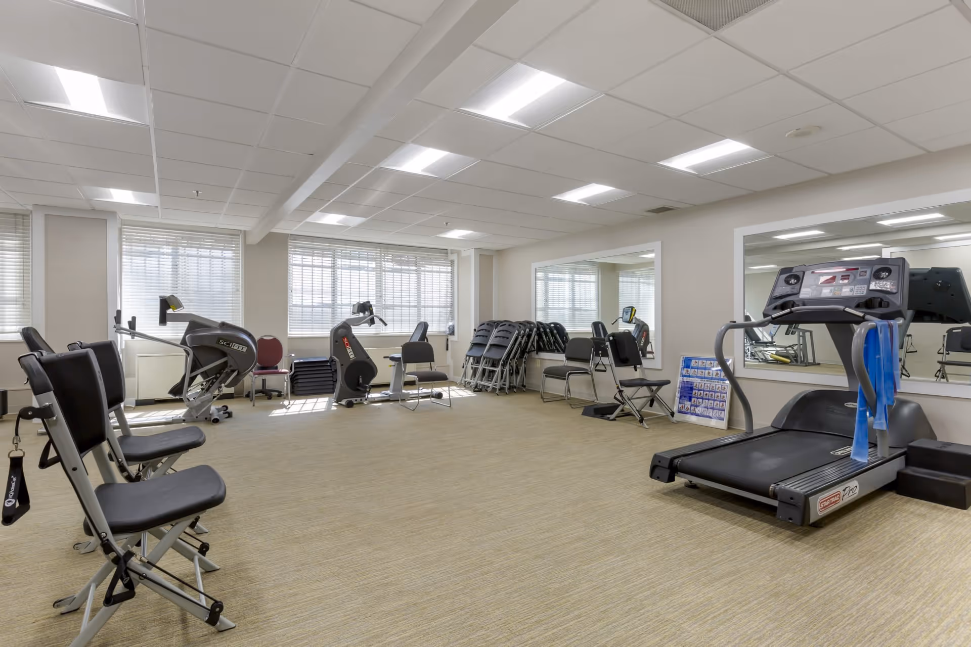A well-lit exercise room with various fitness equipment including a treadmill, stationary bikes, and chairs arranged along the walls. Large windows with blinds allow natural light to fill the room, and a large mirror is mounted on one wall. The floor is carpeted and the ceiling has recessed lighting.