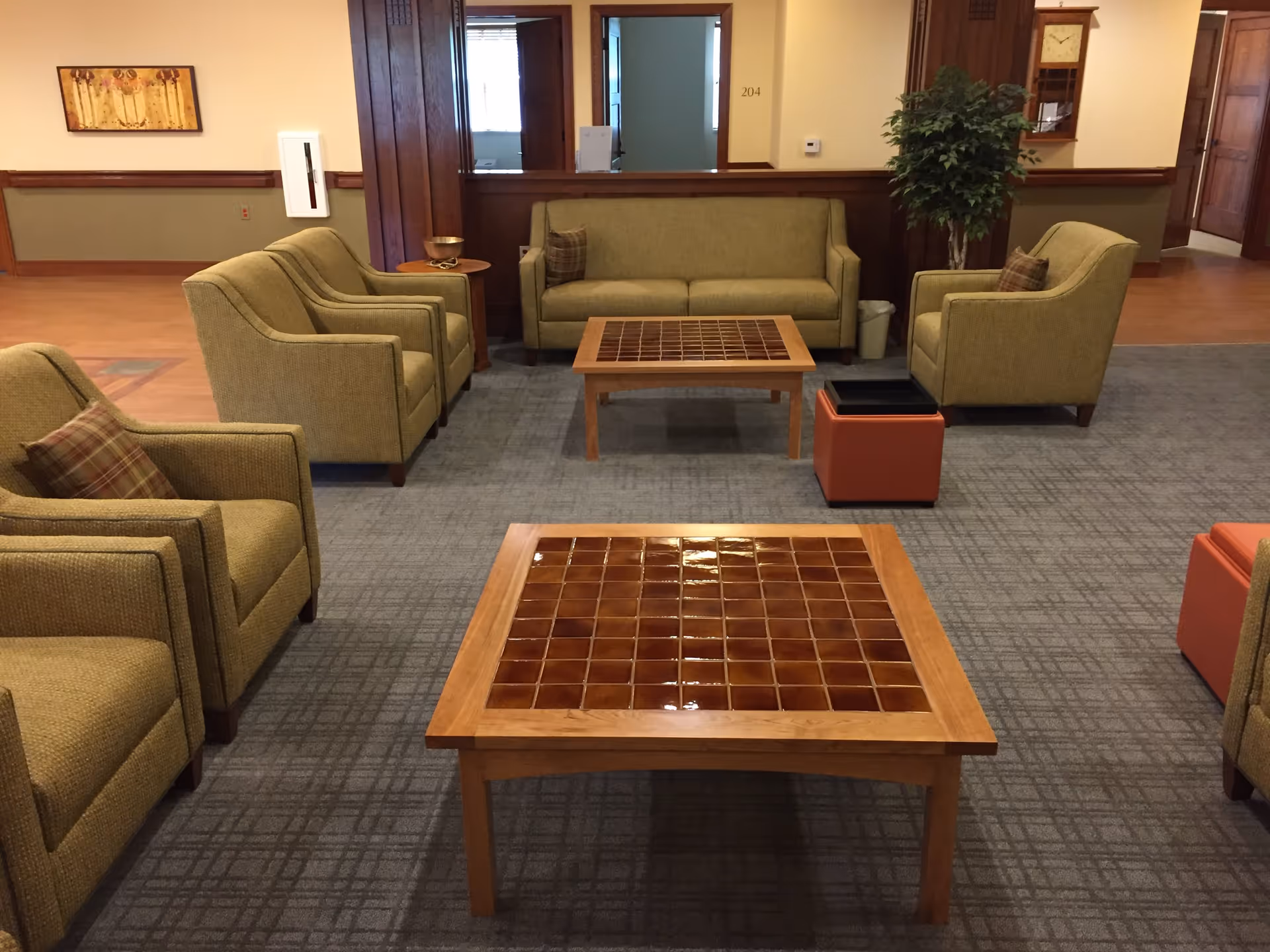A senior living facility common area with green upholstered armchairs and a matching sofa arranged around two wooden coffee tables with tiled tops. There are small side tables, a potted plant, and a wall clock in the background. The floor is carpeted with a patterned design.