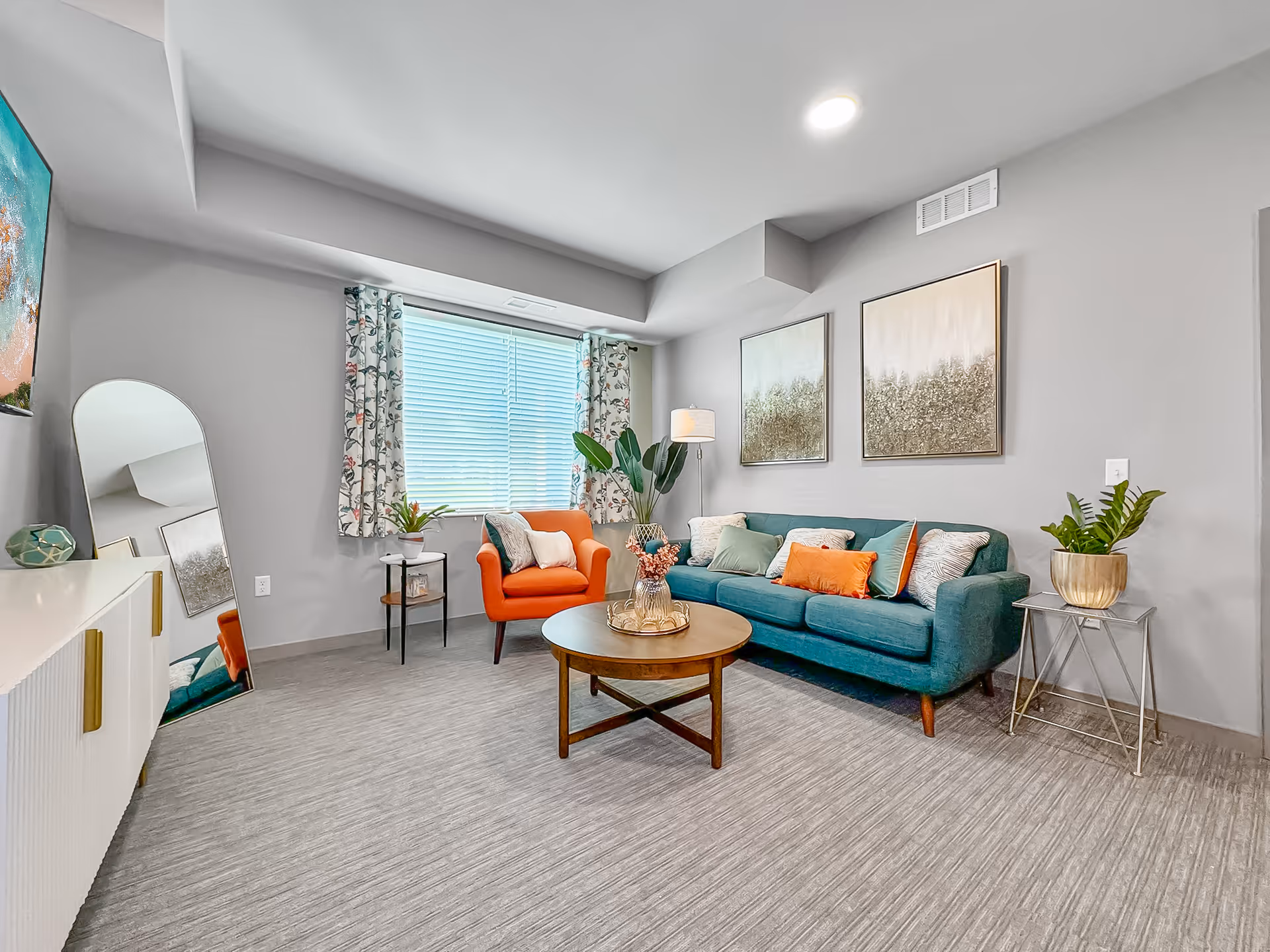 A bright and modern living room with a teal sofa adorned with multiple cushions, an orange armchair, a round wooden coffee table with a decorative centerpiece, a tall floor lamp, two framed artworks on the wall, a large window with floral curtains, a full-length mirror leaning against the wall, and two side tables with potted plants.