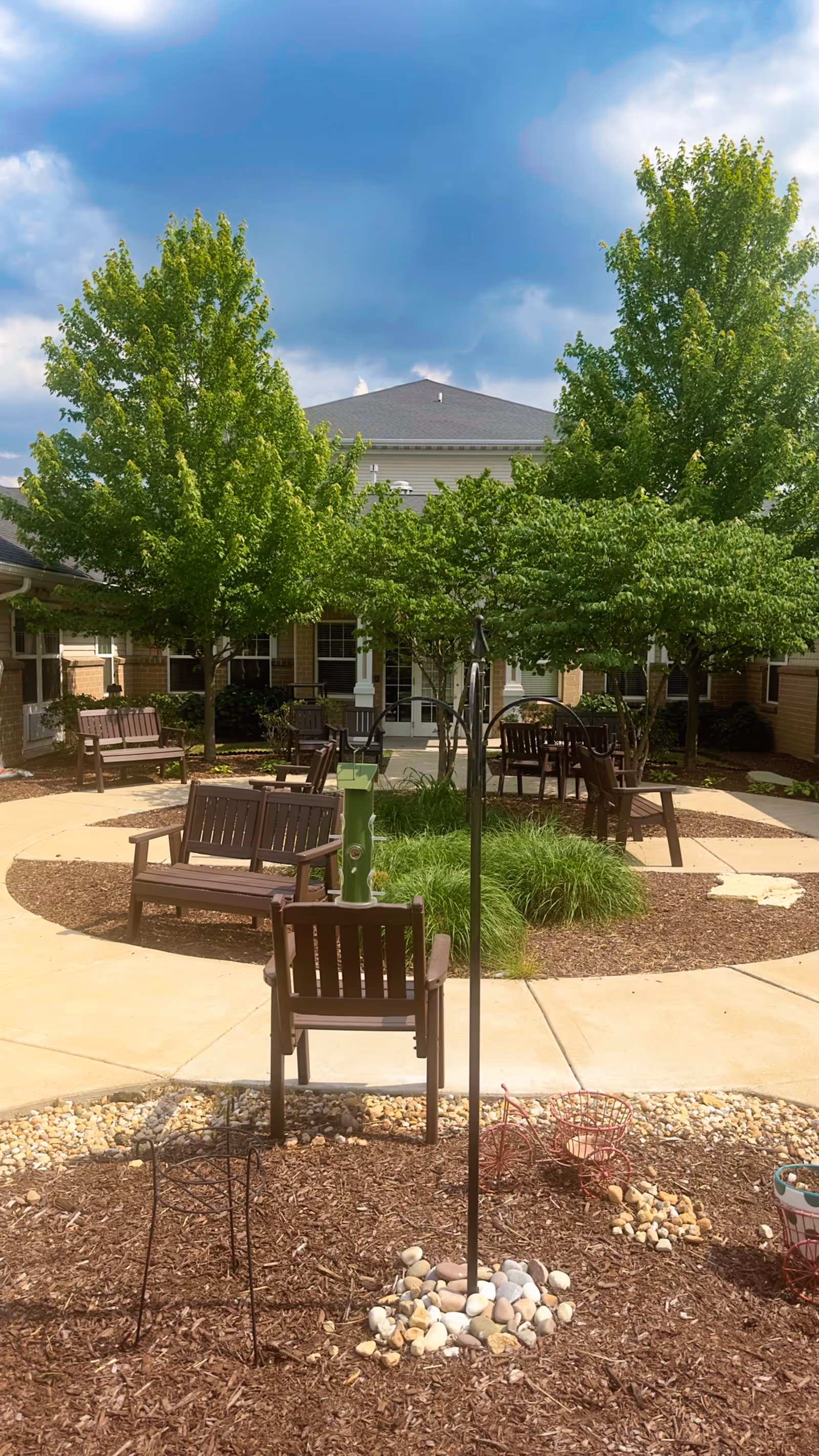 Outdoor courtyard area with several wooden benches and chairs arranged on a circular paved walkway surrounded by mulch and small rocks. There are green trees providing shade and a building with a gray roof in the background under a partly cloudy sky.