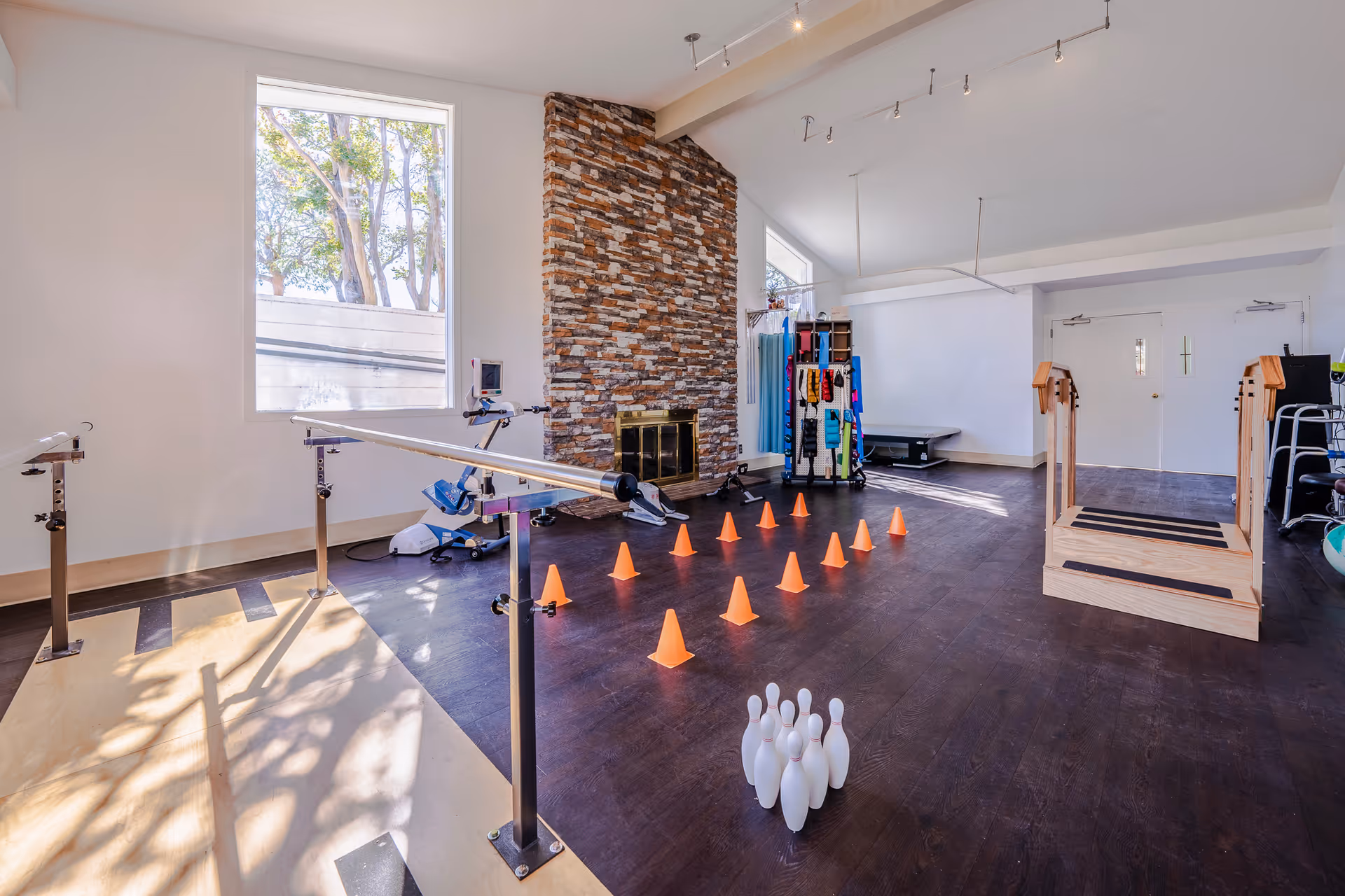Bright rehabilitation/exercise room with parallel bars, orange cones, therapy equipment, bowling pins and a stone fireplace.
