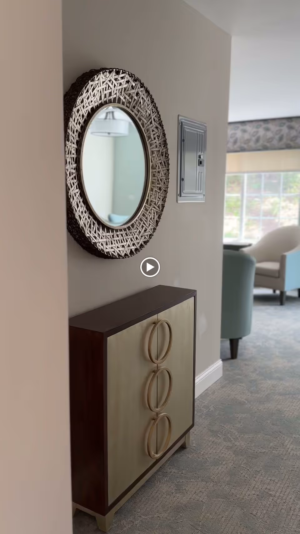 A decorative round mirror with a woven frame hangs on a beige wall above a small cabinet with three large circular handles. In the background, there is a window letting in natural light and a light blue armchair near a patterned wallpaper border.
