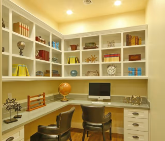 Well-lit study nook with built-in shelves, a corner desk holding a computer and globe, and two leather chairs.