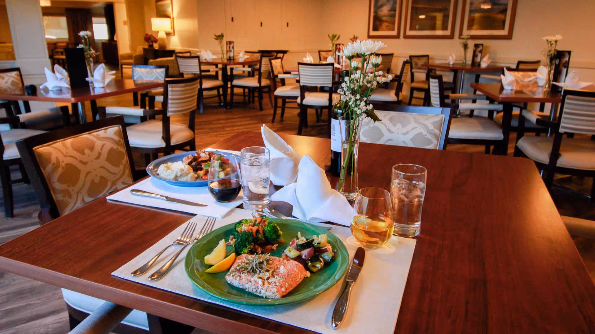 A dining room with multiple tables and chairs. The foreground table is set with two plates of food, glasses of water, a glass of wine, and a small vase with white flowers. The room has warm lighting and framed artwork on the walls.