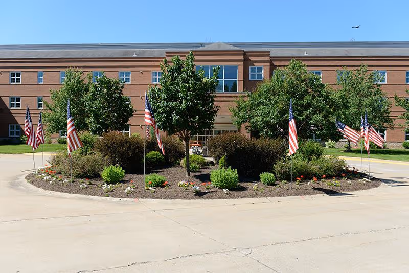 Front exterior view of a large brick building with multiple windows, surrounded by a circular landscaped area with trees, bushes, flowers, and several American flags. A clear blue sky and a small airplane flying in the distance are visible.