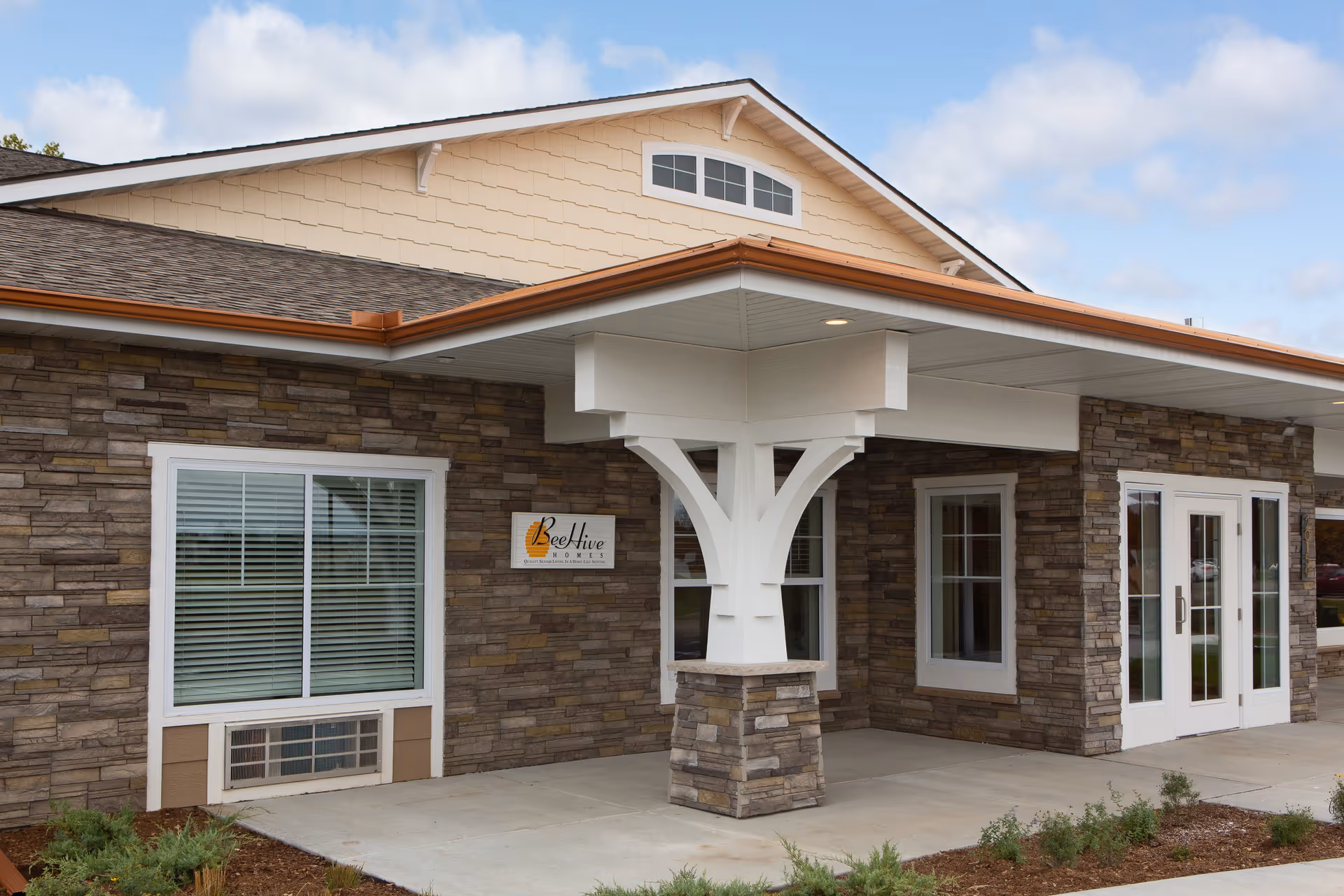 Front entrance of a Beehive Homes building with a stone facade, covered portico, windows and glass double doors.