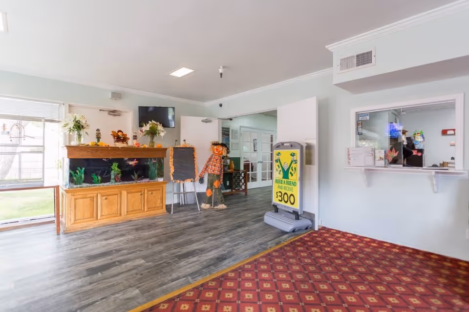 Interior view of a senior living facility lobby with a fish tank on a wooden cabinet, a scarecrow decoration, a chalkboard, a sign advertising a $300 referral reward, a reception window, and a red patterned carpet on the floor.