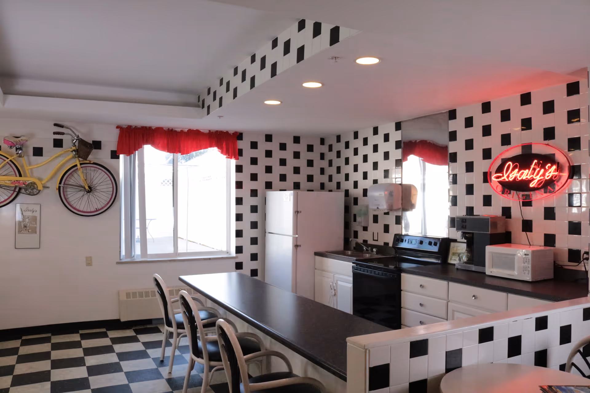 A kitchen area with black and white checkered tile walls and floor. The kitchen includes a white refrigerator, black stove, microwave, coffee maker, and a long black countertop with chairs. A yellow bicycle is mounted on the wall near a window with a red valance. A red neon sign reading 'Italy's' is also visible on the tiled wall.