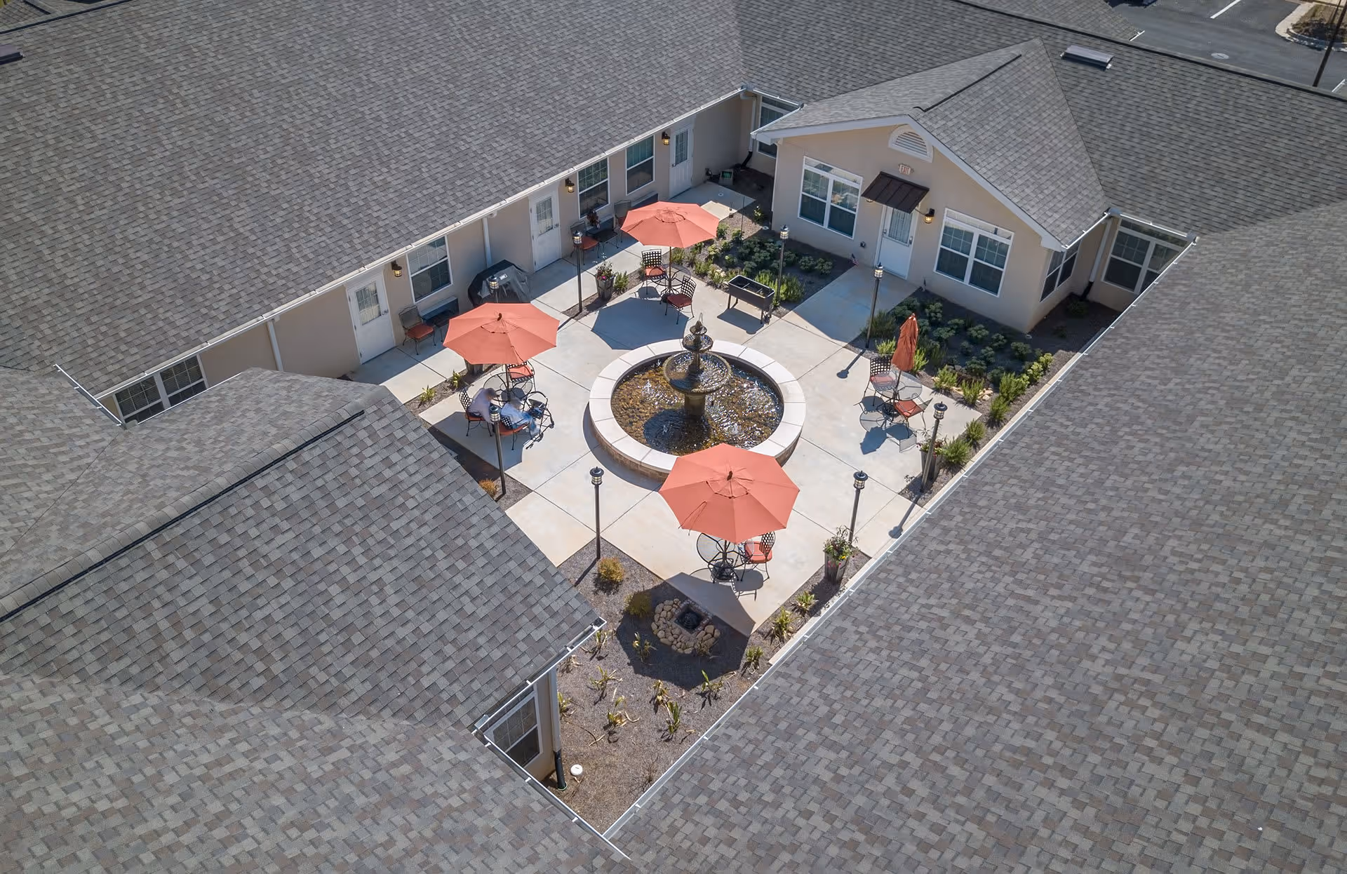 Aerial view of an outdoor courtyard at Benton House of Decatur featuring a central water fountain surrounded by concrete walkways, red patio umbrellas, metal chairs, and small tables. The courtyard is enclosed by single-story buildings with gray shingled roofs and beige walls, with some landscaping along the edges.