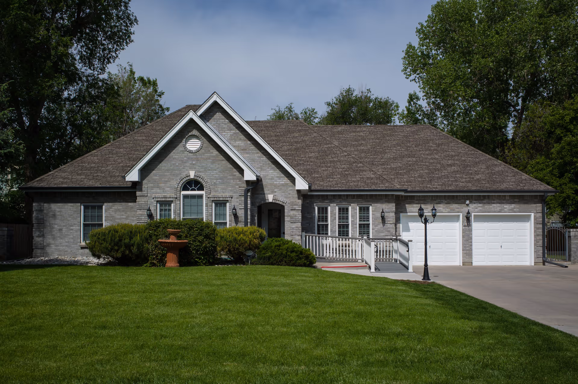 A single-story brick building with a gabled roof, white-framed windows, and a double garage. The building is surrounded by green grass, bushes, and trees, with a concrete driveway and a black lamp post near the entrance.
