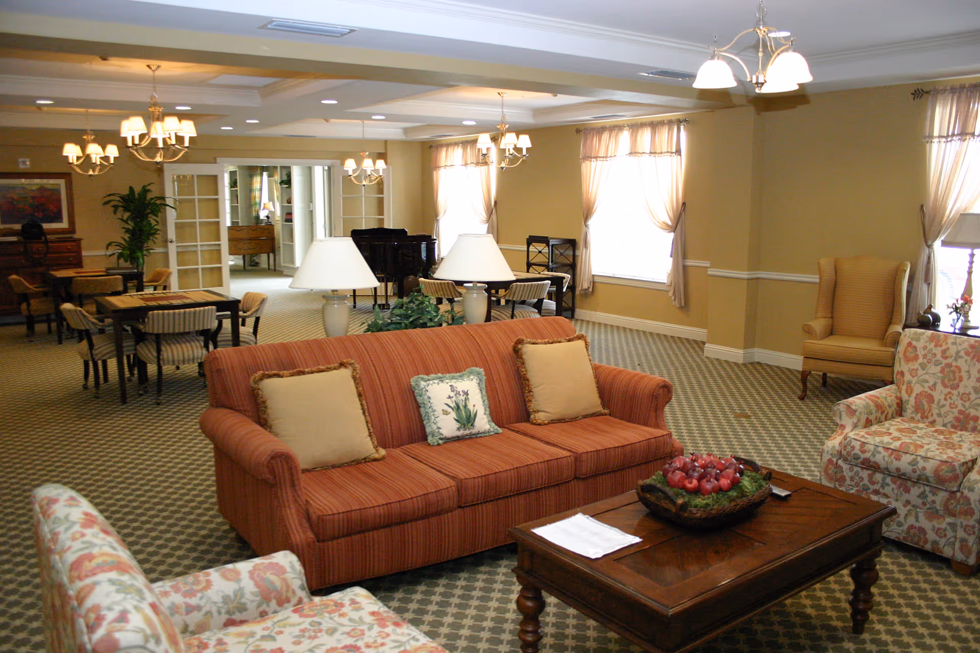 A spacious and well-lit living room area in Prestonwood Court featuring a striped red sofa with three cushions, two floral armchairs, a wooden coffee table with a decorative bowl of red apples, and two table lamps. The room has beige walls, patterned carpet, multiple windows with sheer curtains, chandeliers, and a dining area with tables and chairs in the background.