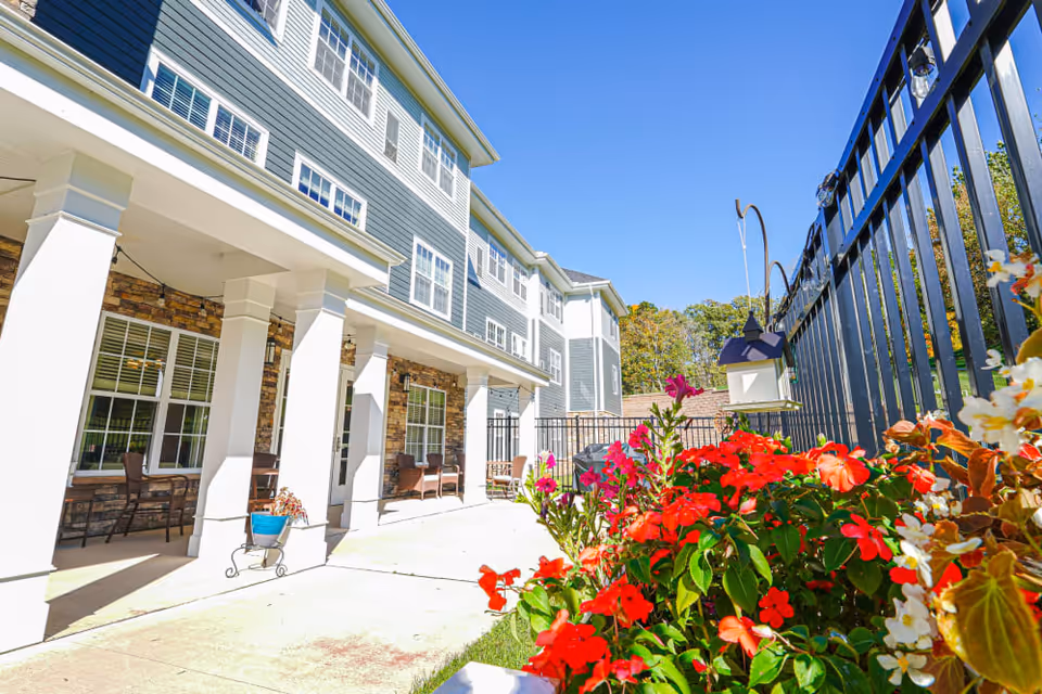 Outdoor patio area of a senior living facility with white columns supporting a covered walkway, several chairs arranged along the building, a black metal fence, and vibrant red and white flowers in the foreground under a clear blue sky.