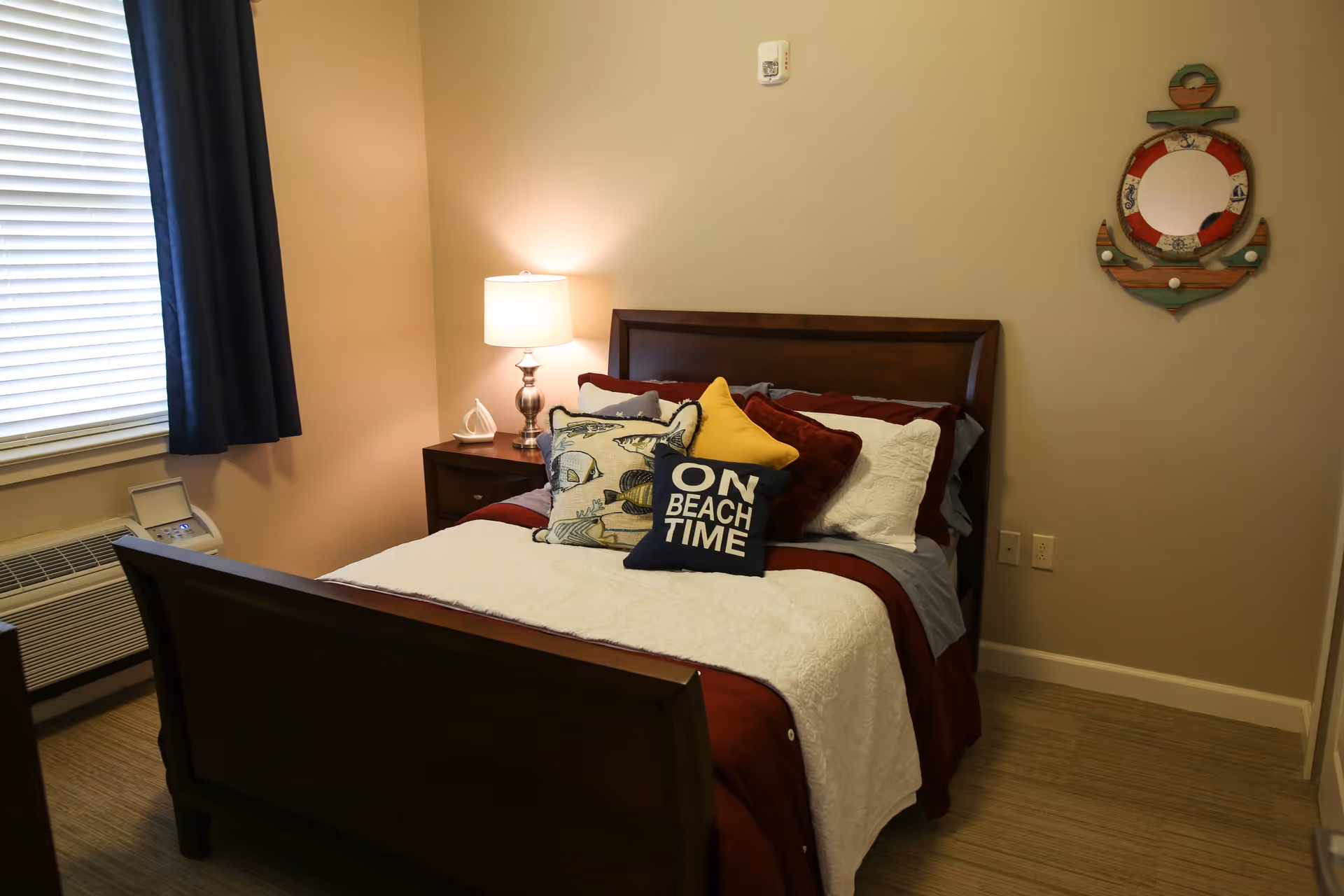 A cozy bedroom with a wooden bed frame, neatly made bed with multiple pillows including one that says 'ON BEACH TIME'. A nightstand with a lamp and a decorative item is beside the bed. There is a window with closed blinds and dark curtains, an air conditioning unit below the window, and a nautical-themed wall decoration shaped like an anchor with a circular mirror in the center.