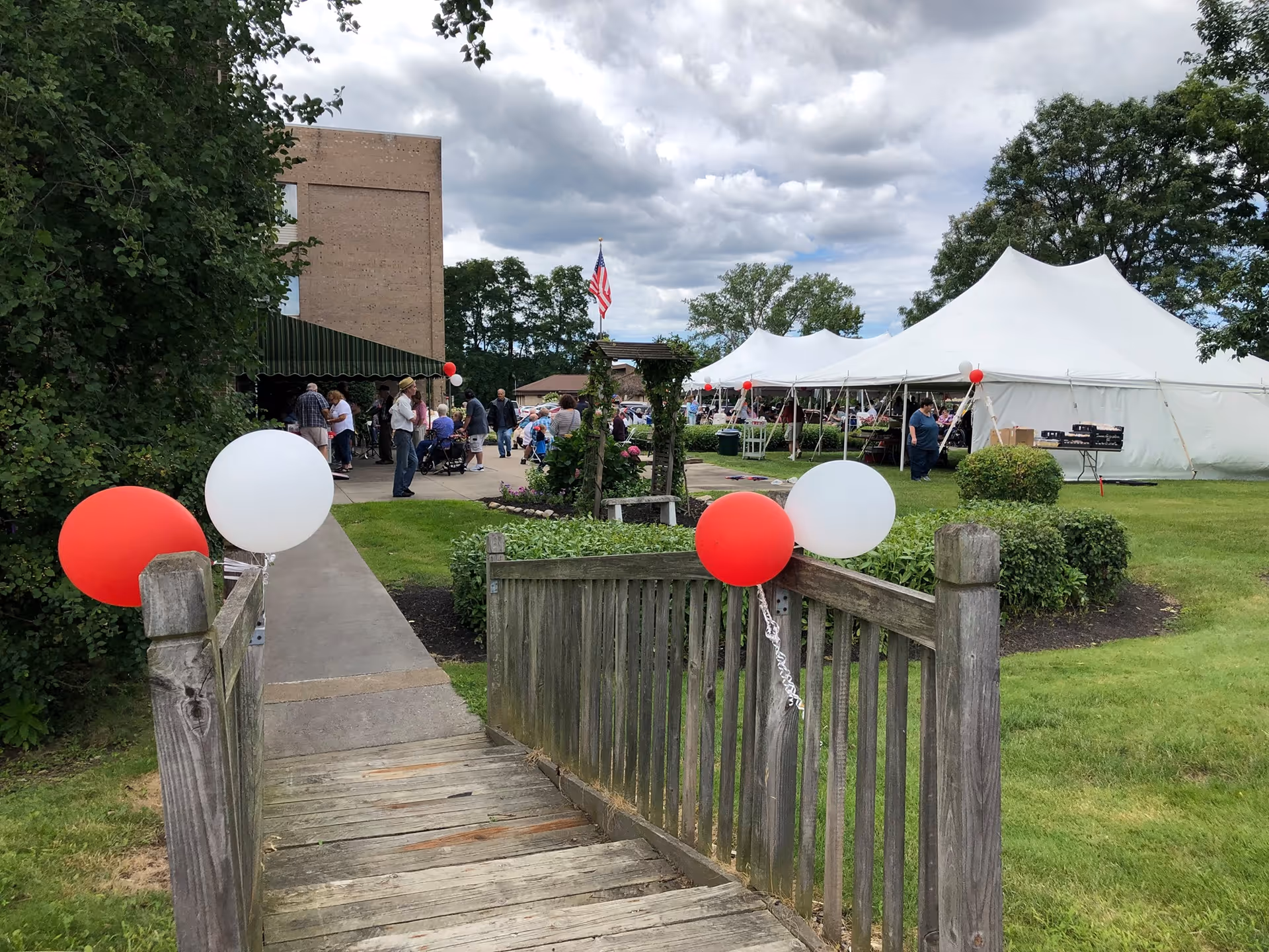 Outdoor view of a walkway and wooden bridge decorated with red and white balloons leading to tents and a building where people are gathered on a lawn.