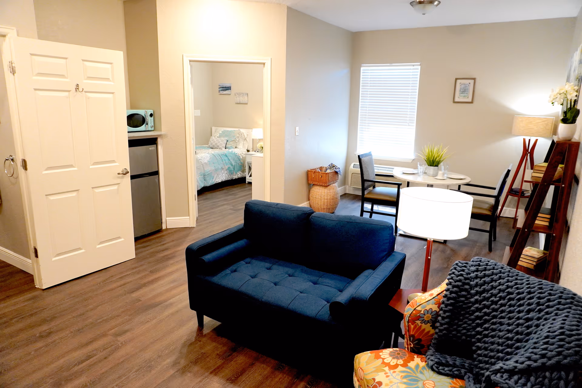 A cozy living area with a dark blue loveseat and a colorful armchair with a dark knitted throw. There is a small round dining table with two chairs near a window with blinds. A wooden ladder shelf with books and a lamp is against the wall. In the background, an open door reveals a bedroom with a bed covered in light blue bedding. A small kitchenette area with a mini fridge and microwave is visible near the bedroom door. The room has wood flooring and neutral-colored walls.