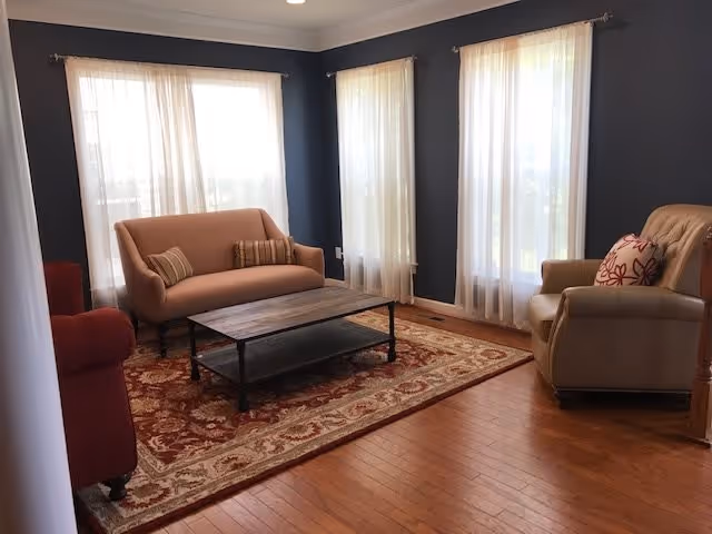 A cozy living room with dark blue walls and three large windows covered with sheer white curtains. The room features a beige loveseat with two striped cushions, a beige armchair with a floral cushion, a red armchair, a wooden coffee table with a metal frame, and a large patterned area rug on a wooden floor.