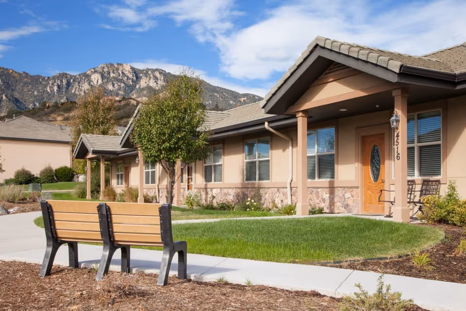 Exterior view of a single-story residential building with beige walls, stone accents, and a wooden front door with the number 4516. There is a small covered porch with two chairs, a green lawn, a tree, and a wooden bench on a paved walkway. Mountains and a partly cloudy sky are visible in the background.