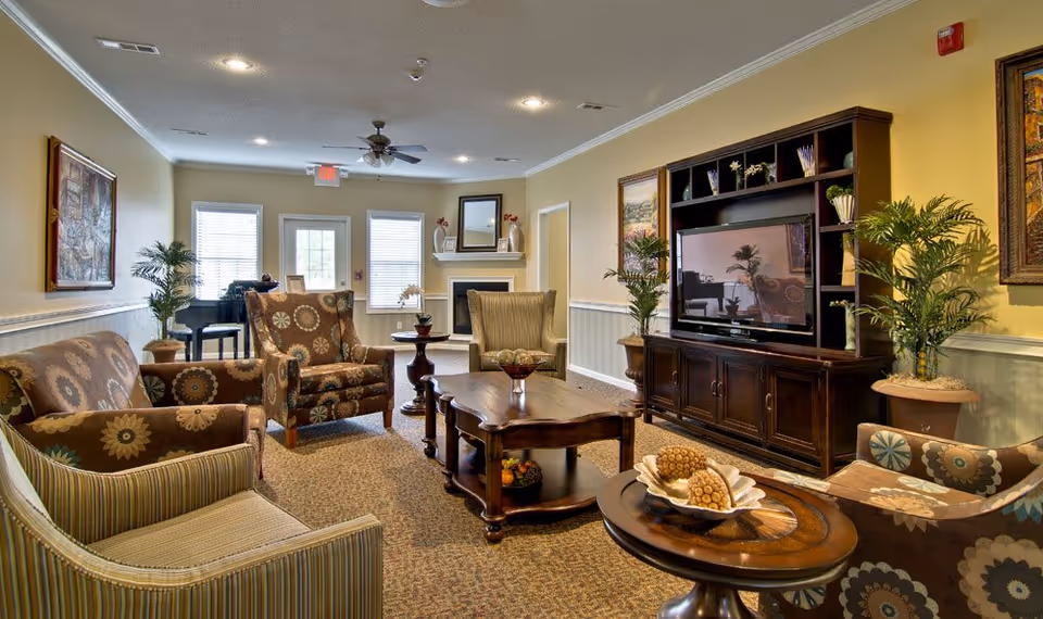 A cozy living room with patterned armchairs and a sofa arranged around a wooden coffee table. There is a large dark wood entertainment center with a flat-screen TV, decorative plants, and framed artwork on the walls. The room has beige walls, carpeted floor, and a ceiling fan with lights. Windows and a door are visible at the far end, letting in natural light.