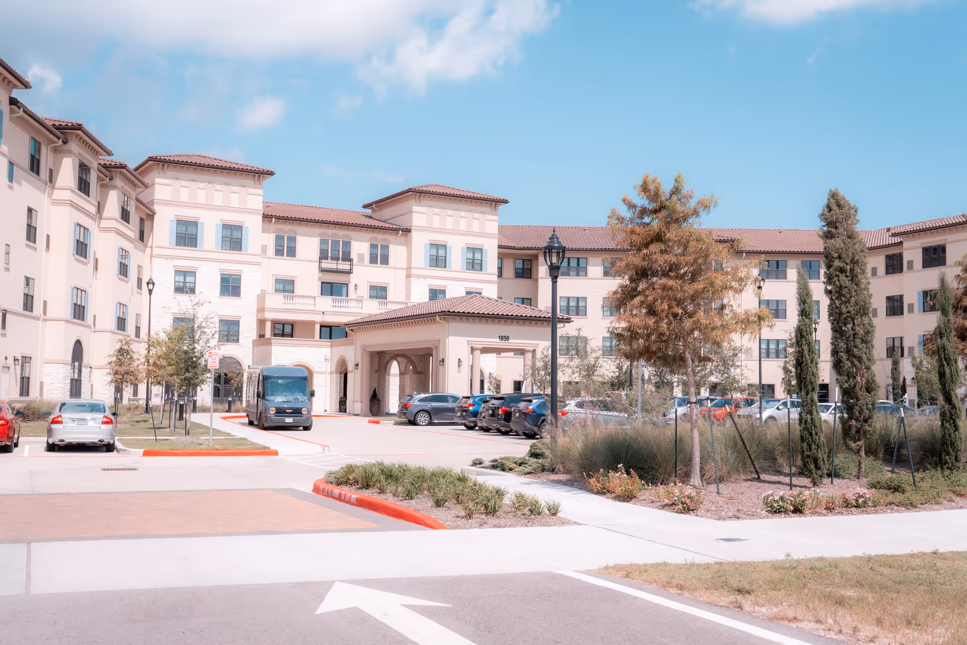 Exterior view of a large multi-story senior living facility building with beige walls and red-tiled roofs under a blue sky. There is a driveway with parked cars and a shuttle bus near the entrance, surrounded by landscaped greenery and trees.