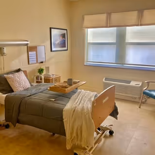 A bright senior bedroom with an adjustable hospital-style bed, bedside tables, a window with blinds, an armchair and wall art.