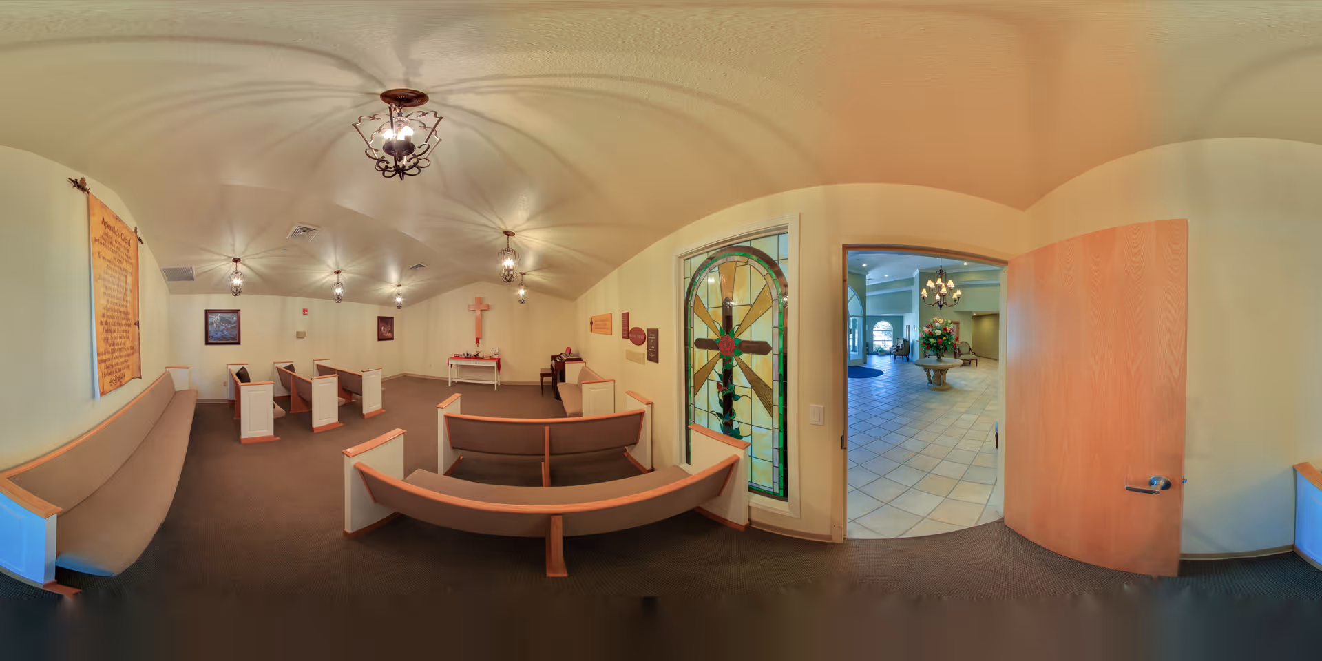 Interior view of a small chapel room with wooden pews arranged facing a wall with a cross and a small altar. The room has beige walls, carpeted floor, and decorative hanging light fixtures. A stained glass window with a cross design is visible next to an open wooden door leading to a tiled lobby area with a round table and floral arrangement.