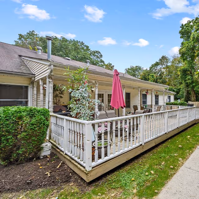 Outdoor view of a single-story building with a wooden deck surrounded by a white railing. The deck has patio furniture including chairs and a table with a closed pink umbrella. There are potted plants on the deck and greenery including bushes and trees around the building under a partly cloudy sky.