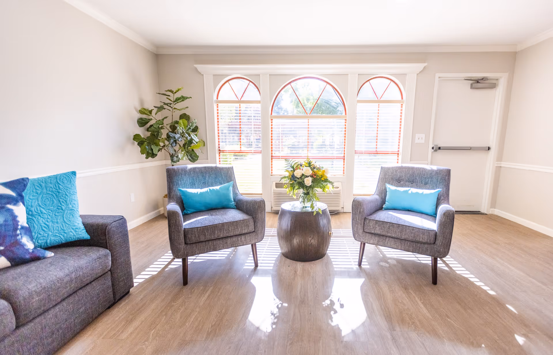 A bright and airy sitting area with two gray armchairs and a gray sofa, each with blue accent pillows. A small round wooden table with a floral arrangement is placed between the armchairs. Large windows with red trim and white blinds allow natural light to fill the room. A green potted plant is in the corner, and the floor is light wood.