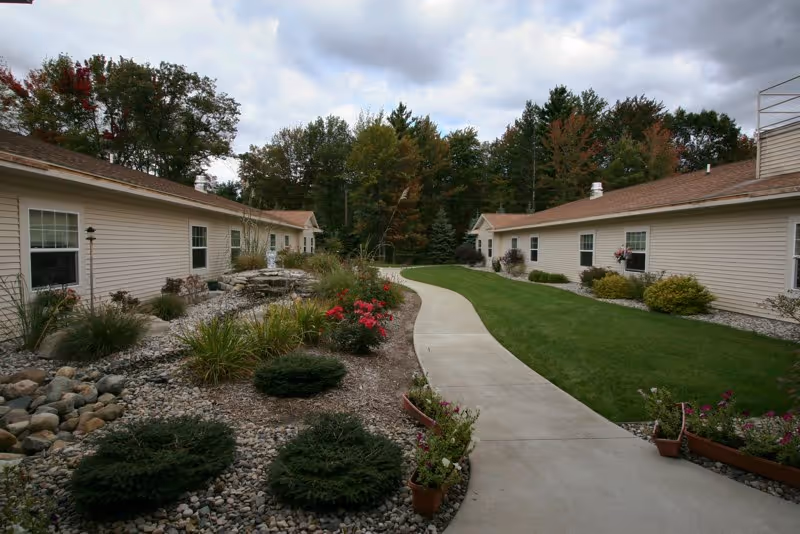 A paved walkway curves through a landscaped outdoor area between two single-story beige buildings with brown roofs. The area features green grass, various shrubs, flowering plants, and small trees, with a backdrop of taller trees under a cloudy sky.
