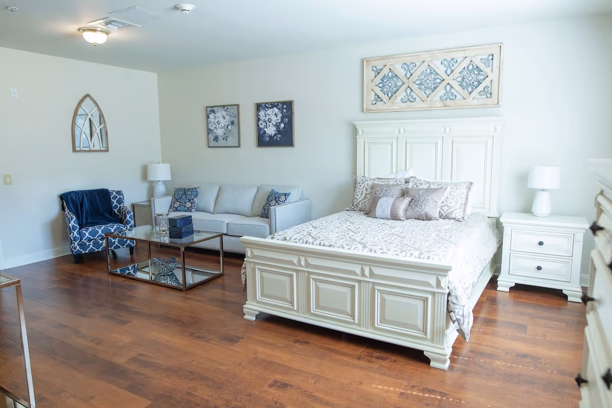 A bright and spacious bedroom featuring a large white wooden bed with decorative pillows and a patterned bedspread. To the right of the bed is a white nightstand with a white lamp. On the left side of the room, there is a light gray sofa with patterned cushions, a glass coffee table with decorative items, and a blue patterned armchair with a dark blue throw blanket. The walls are decorated with floral artwork and a decorative mirror. The floor is wooden with a warm tone.