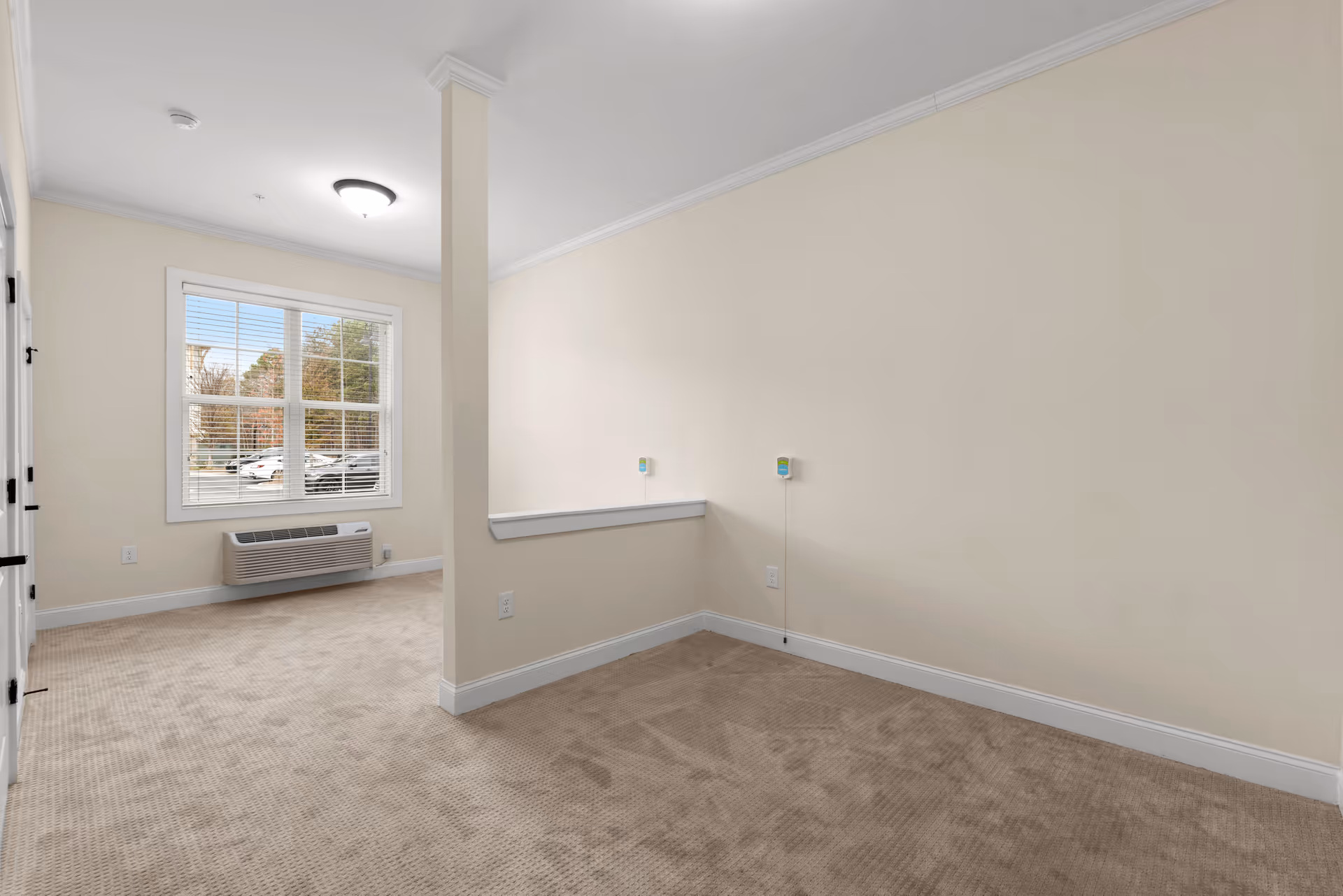 Empty room with beige carpet and cream-colored walls, featuring a window with white blinds showing a view of parked cars and trees outside. The room has a white baseboard, a ceiling light fixture, and two wall-mounted devices with cords on one wall. A partial wall divides the space.