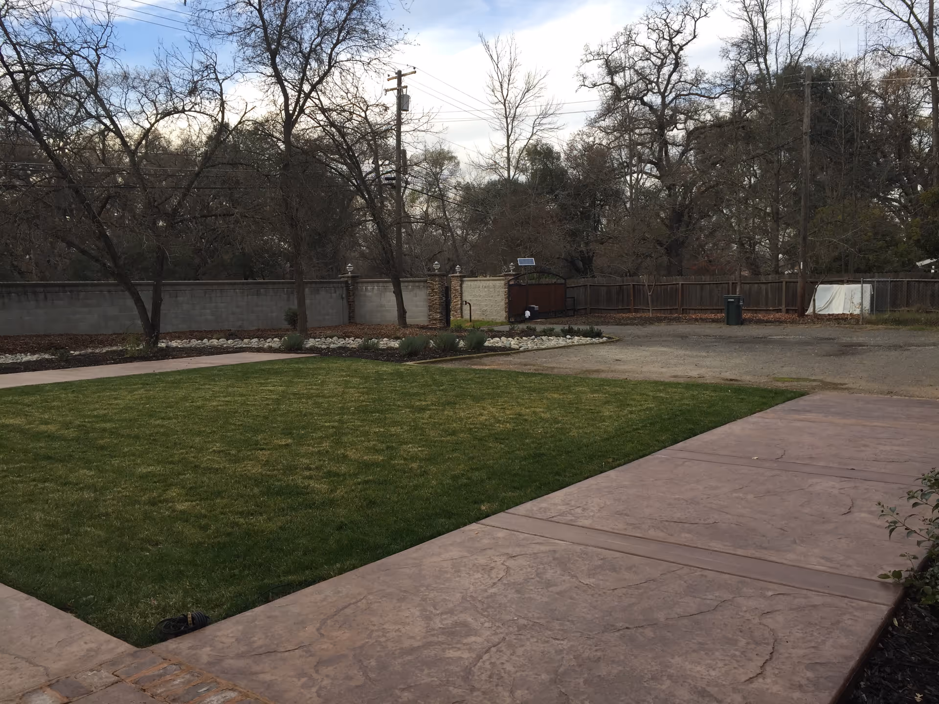 Outdoor area with a green lawn bordered by a paved walkway, surrounded by leafless trees and a wooden fence in the background under a cloudy sky.