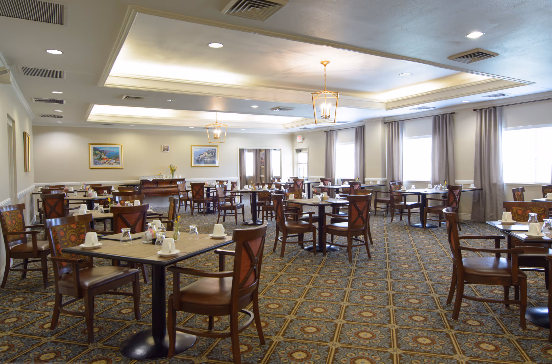 A spacious dining room with multiple tables and chairs arranged neatly. Each table is set with white cups, glasses, and small condiment containers. The room features patterned carpet flooring, light-colored walls with framed artwork, large windows with curtains, and ceiling lights with decorative fixtures.