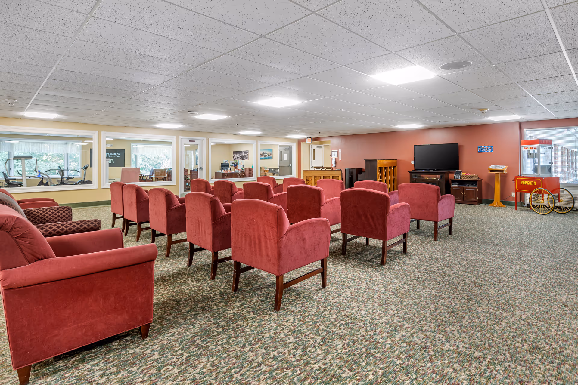 A spacious common area with multiple red upholstered chairs arranged in rows facing a flat-screen TV mounted on a wooden cabinet. There is a popcorn machine on the right side near large windows, and a piano against the far wall. The room has a patterned carpet and a drop ceiling with fluorescent lighting. Through the windows on the left wall, a fitness room with exercise equipment is visible.