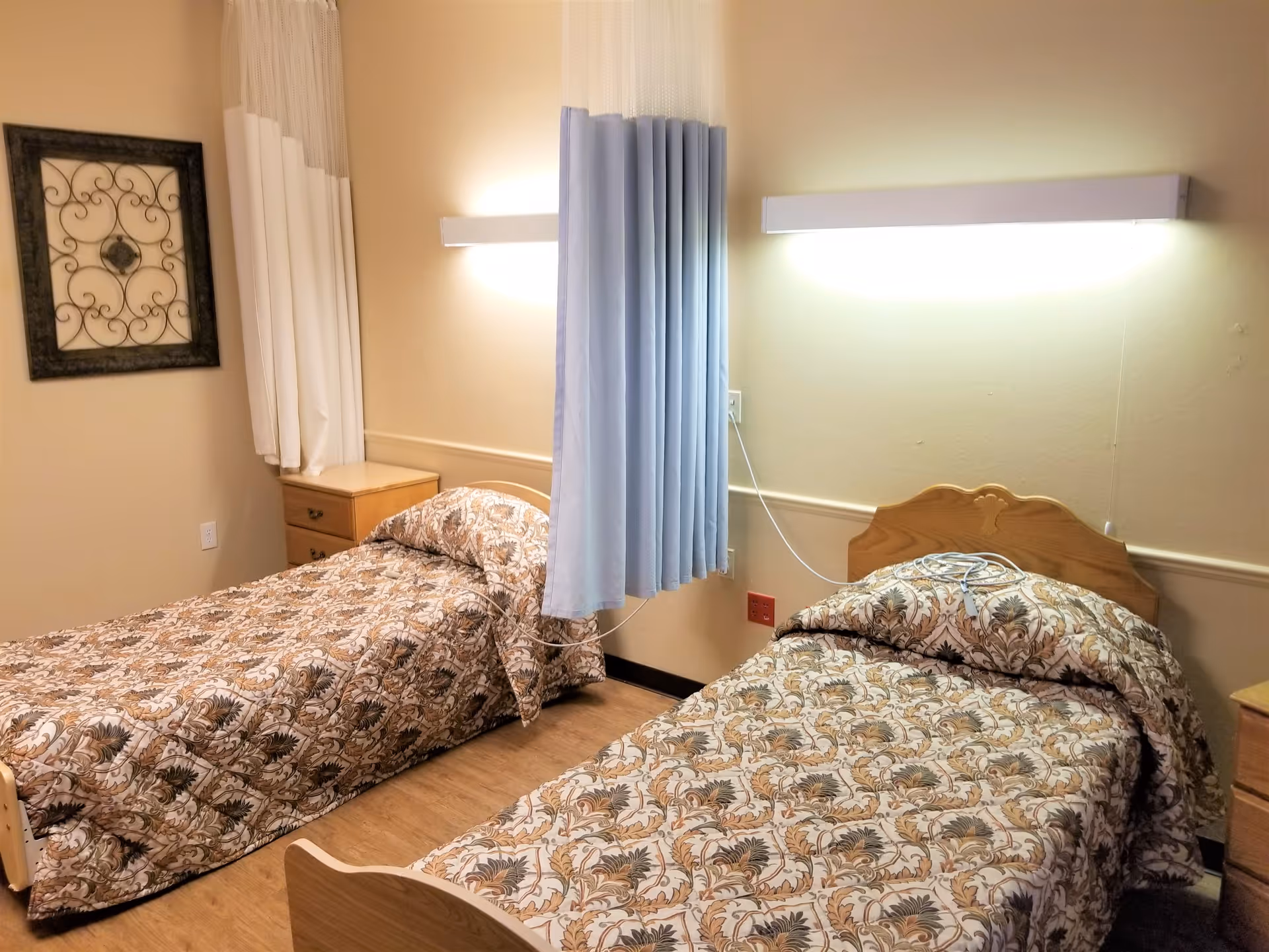 A nursing home bedroom with two single beds separated by a blue privacy curtain. Each bed has a patterned bedspread with floral designs. There are wall-mounted lights above each bed, a wooden nightstand between the beds, and a decorative framed wall art on the left wall.