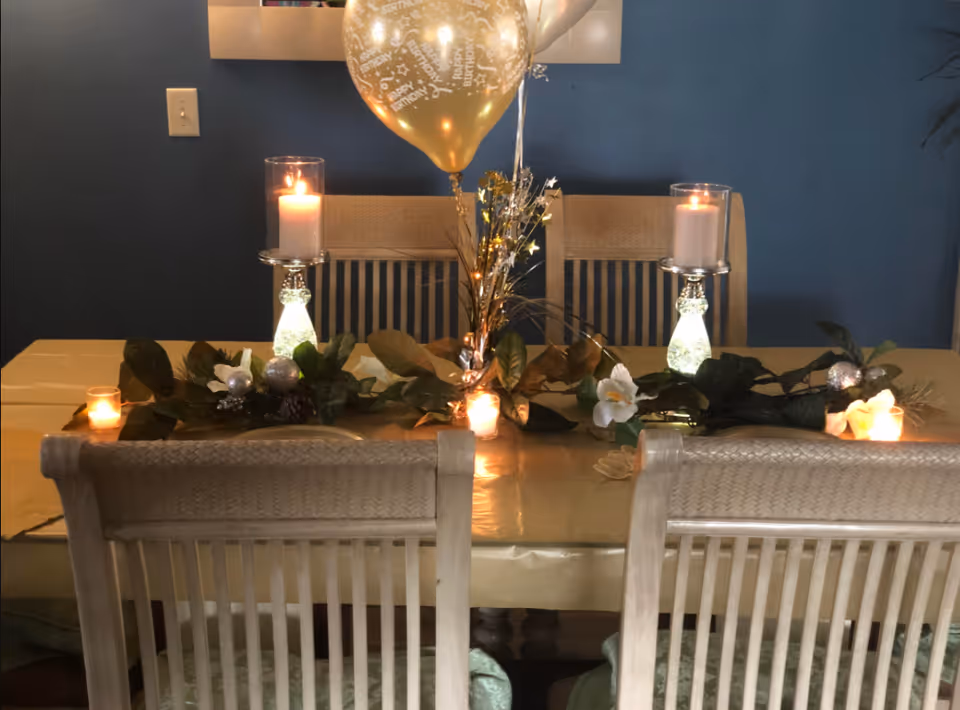 A dining table decorated with a gold tablecloth, green foliage centerpiece, white flowers, two lit candles in glass holders, and gold 'Happy Birthday' balloons against a dark blue wall.