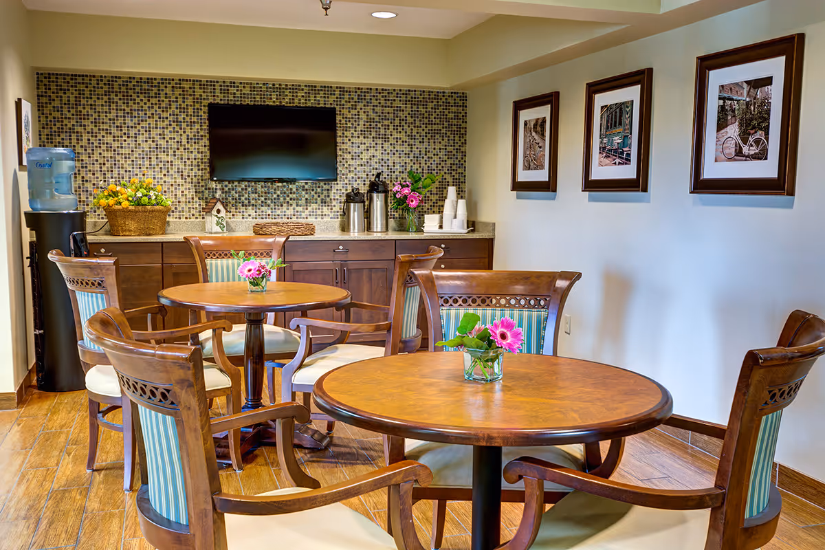 A cozy dining area with two round wooden tables, each surrounded by four wooden chairs with striped upholstery. Each table has a small vase with pink flowers. In the background, there is a countertop with a mosaic tile backsplash, a mounted flat-screen TV, a water dispenser, coffee urns, disposable cups, and decorative items including a basket of yellow flowers and a small birdhouse. Three framed pictures hang on the wall to the right.
