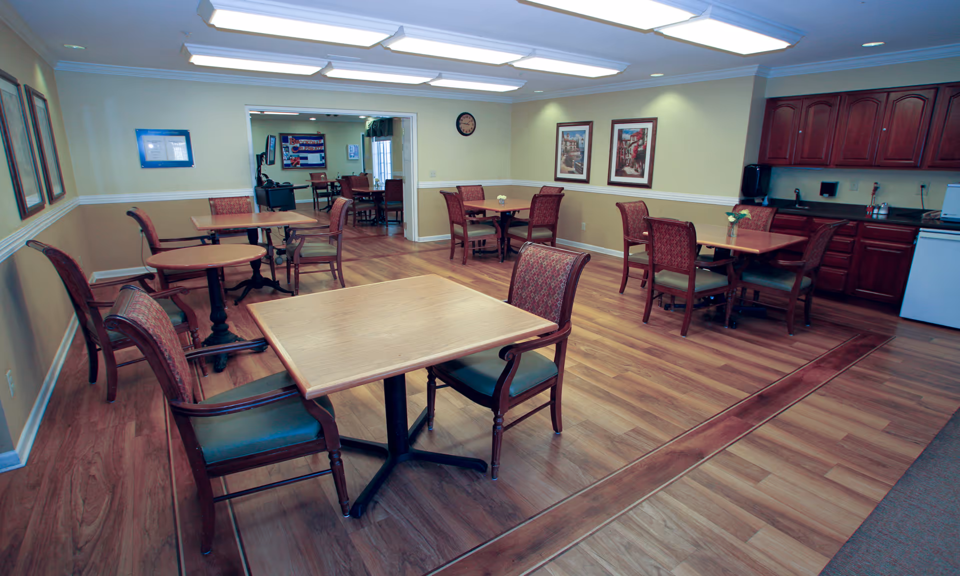 Spacious common dining area with wooden tables and chairs, wood flooring, a kitchenette and framed artwork on the walls.
