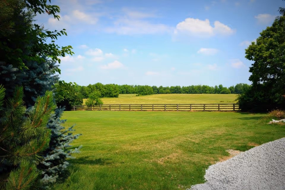 A scenic outdoor view of a large grassy field bordered by a wooden fence, with trees on the left and right sides under a partly cloudy blue sky.