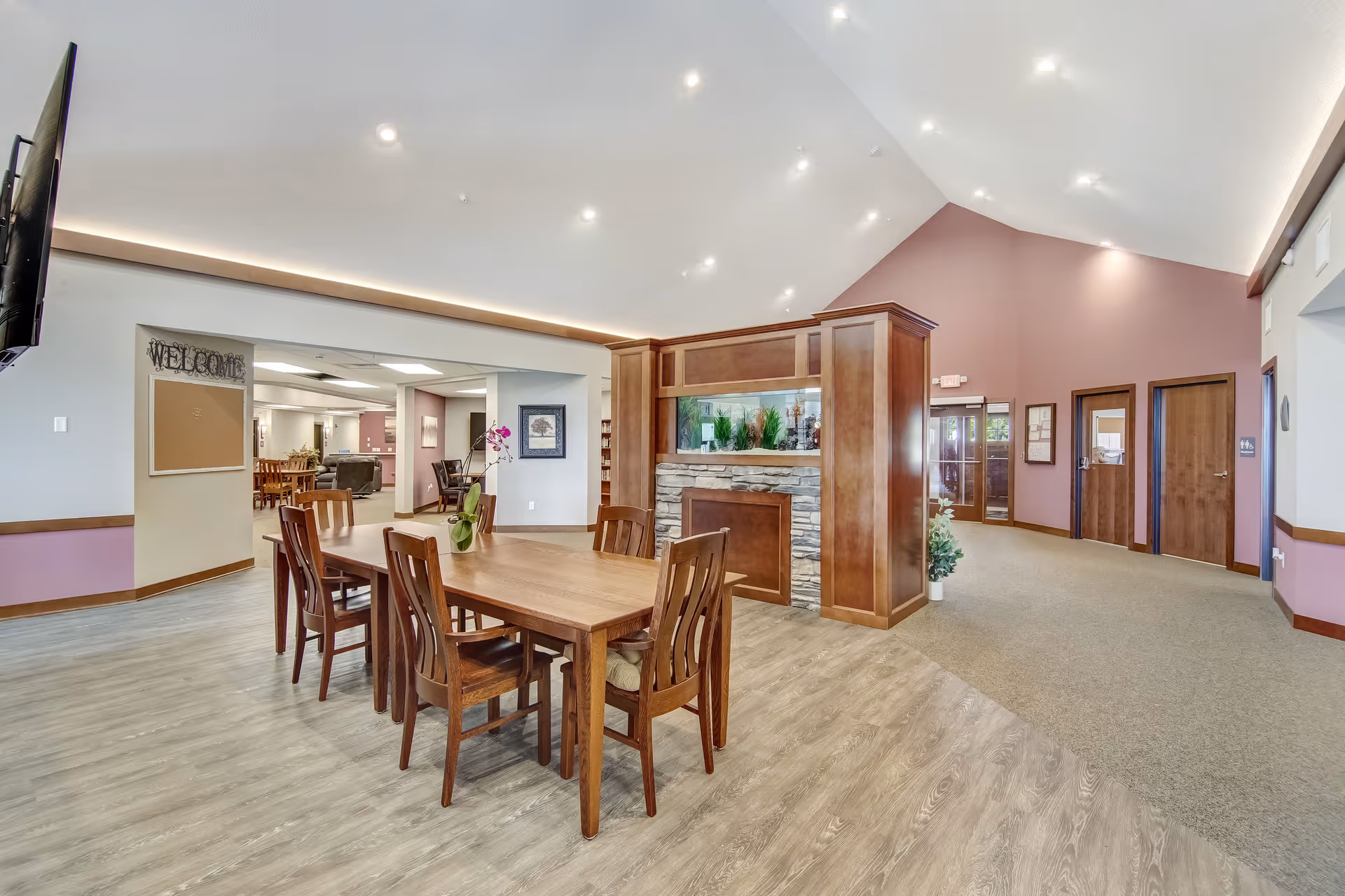 Interior view of a senior living facility common area with a wooden dining table and six chairs in the foreground. The room features a high vaulted ceiling with recessed lighting, a decorative stone and wood partition with an aquarium, and a mix of wood and carpet flooring. There are doors and a hallway leading to other parts of the facility, and a wall-mounted TV is visible on the left side.