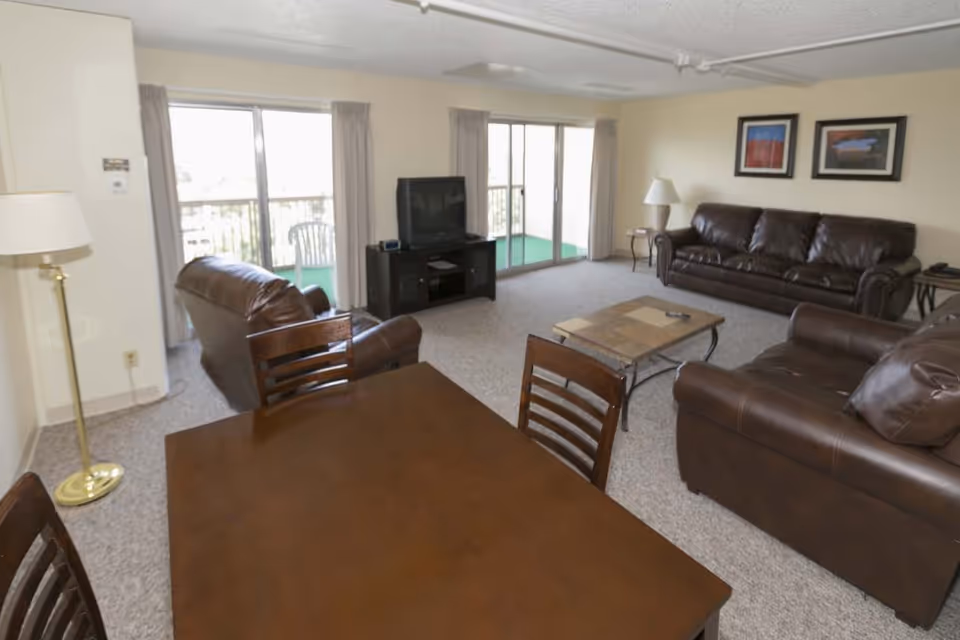 Spacious living room with brown leather sofas, a coffee table, a TV on a stand, and a dining table near sliding glass doors to a balcony.