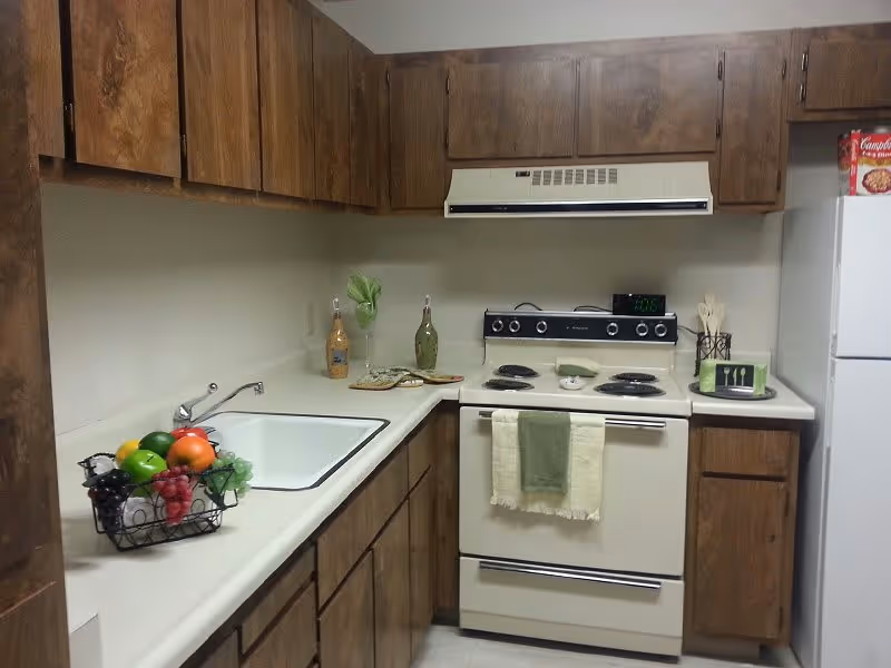 A kitchen with wooden cabinets, a white countertop, a white sink, and a white electric stove with an oven. There is a green and beige towel hanging on the oven door. On the countertop, there are decorative bottles, a basket of artificial fruit, a clock on the stove, and a white refrigerator with a box of Campbell's soup on top.