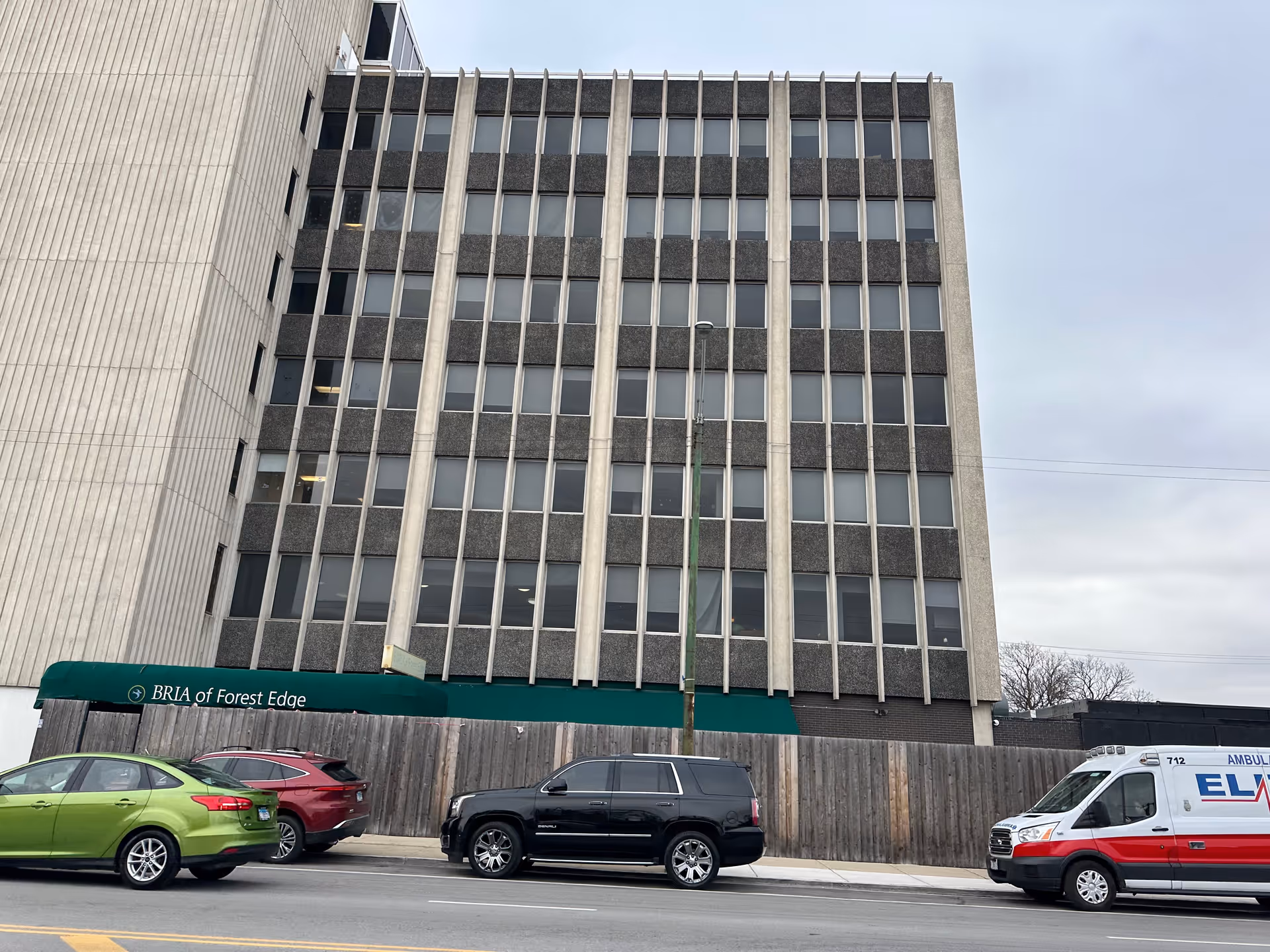Exterior view of a multi-story building with a green awning that reads 'BRIA of Forest Edge'. Several cars and an ambulance are parked along the street in front of the building. The sky is overcast.