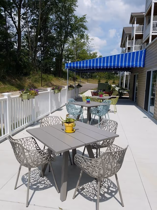 Outdoor patio area with modern tables and chairs arranged on a concrete surface. Each table has a small potted plant. There is a white fence with flower boxes along the edge and a blue and white striped awning providing shade. Trees and greenery are visible in the background.