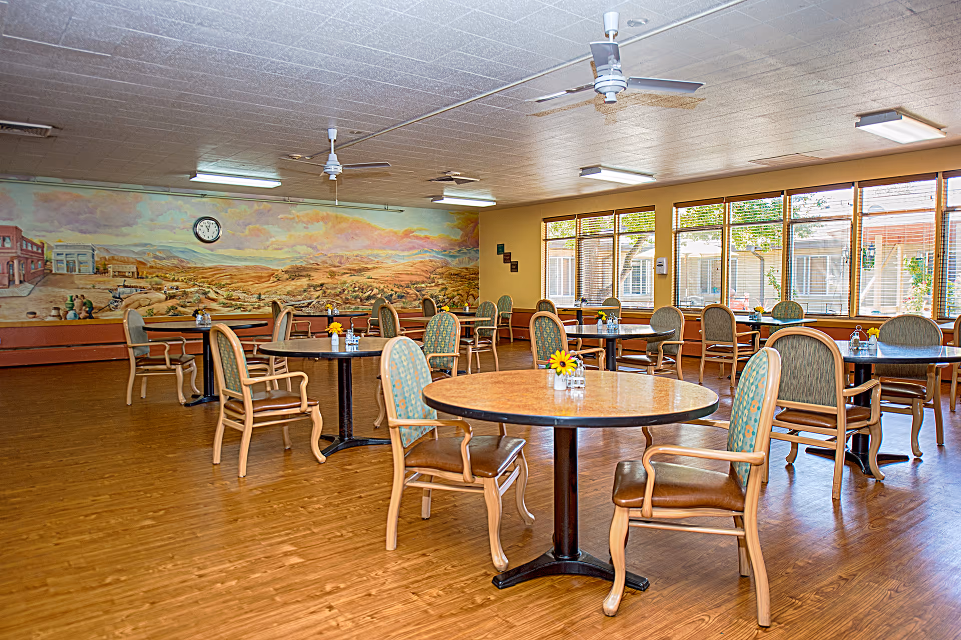 A spacious dining room with multiple round tables, each surrounded by wooden chairs with cushioned seats and backs. The room has large windows letting in natural light, a mural painting on one wall, ceiling fans, and a clock above the mural. Small flower arrangements are placed on the tables.
