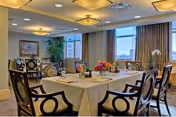 A well-lit dining room with a table set for six people, featuring white tablecloths, floral centerpieces, and elegant chairs. Large windows with curtains allow natural light to fill the room, and there is a framed picture and a tall plant in the background.