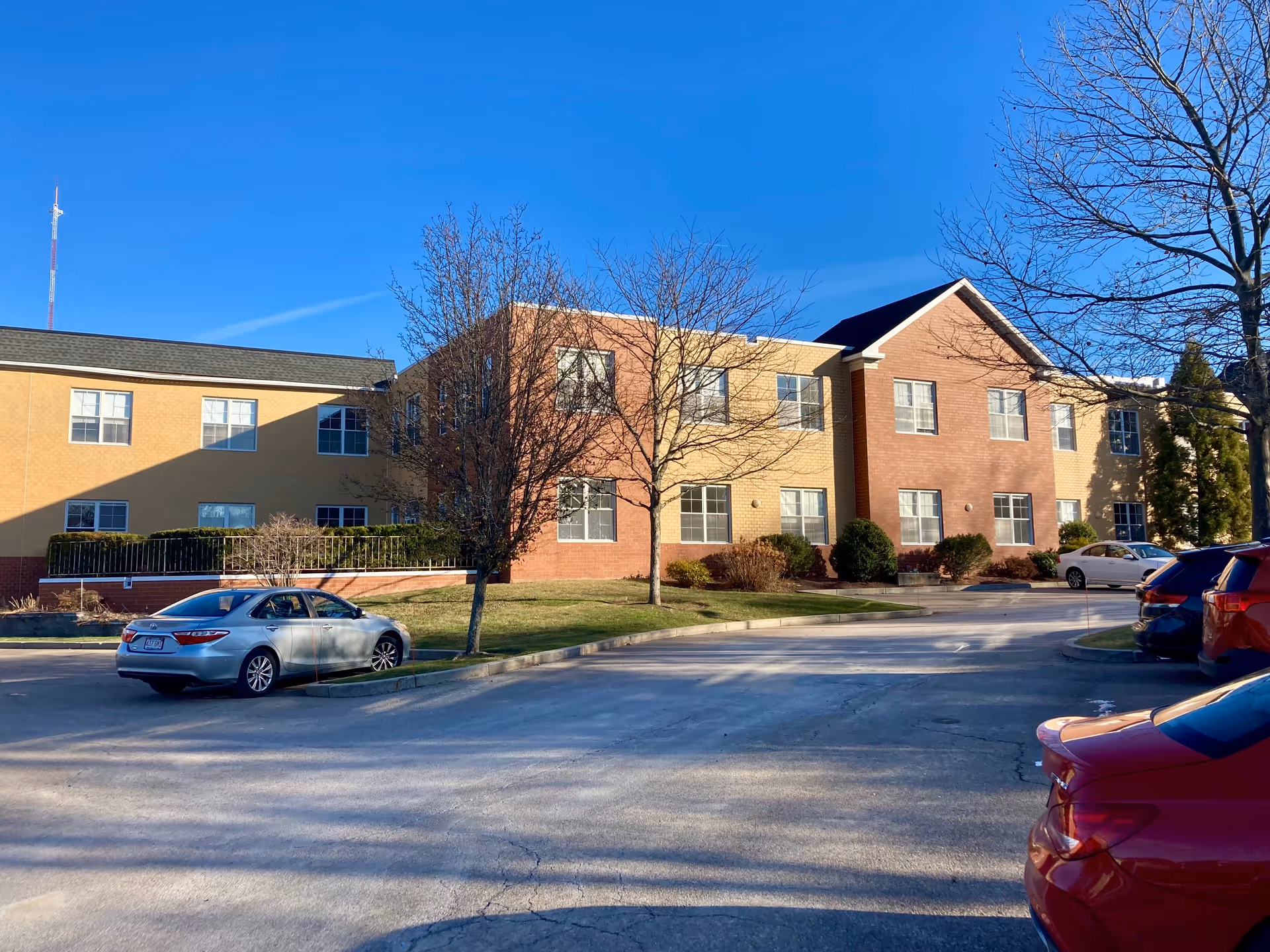 Exterior view of a two-story brick-and-beige building with leafless trees and several parked cars in a front parking lot under a clear blue sky.