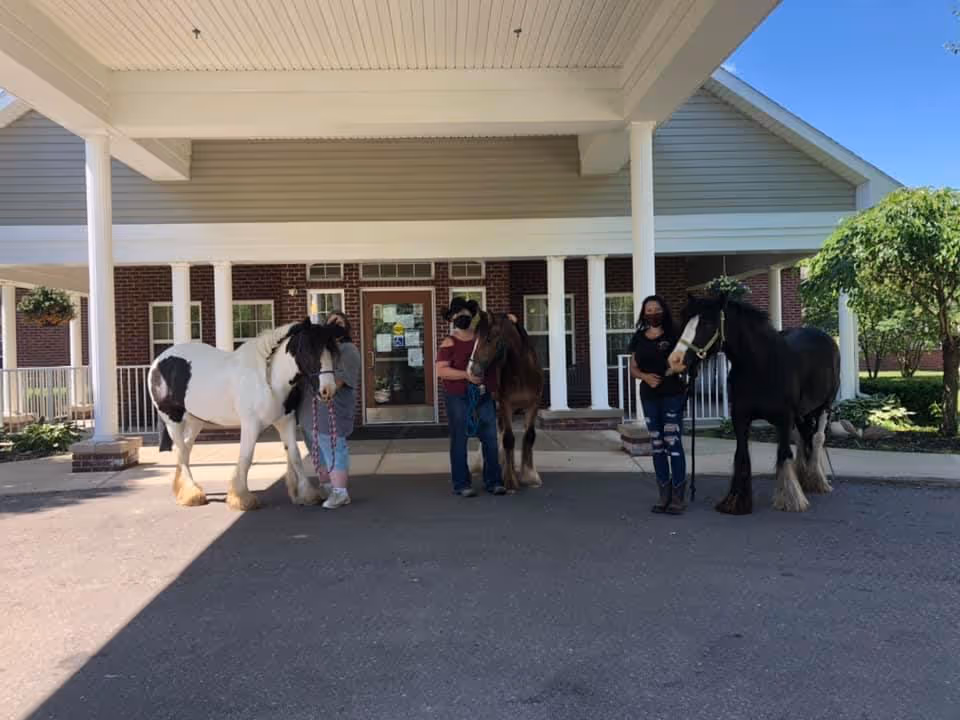 Three people wearing masks stand under a covered entrance holding three horses in front of a brick senior living facility.