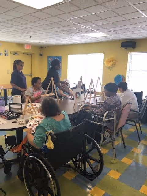 A group of elderly individuals, some in wheelchairs, seated around a table in a brightly lit room engaged in a painting activity with easels and art supplies. Two staff members are standing nearby assisting them. The room has yellow walls and large windows letting in natural light.